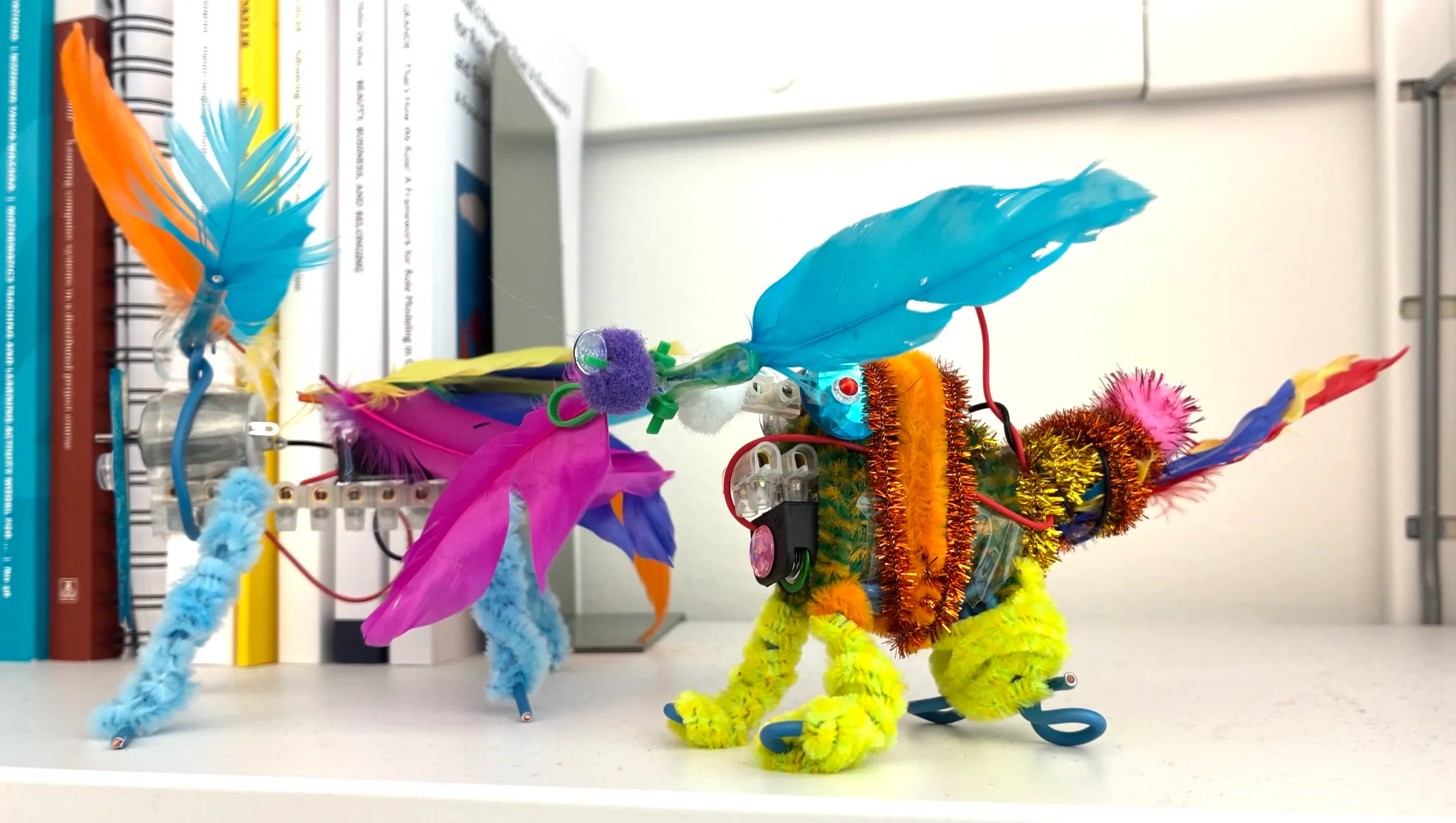 Two colourful bibberich vibrobots on a shelf, built from small motors, wires and feathers